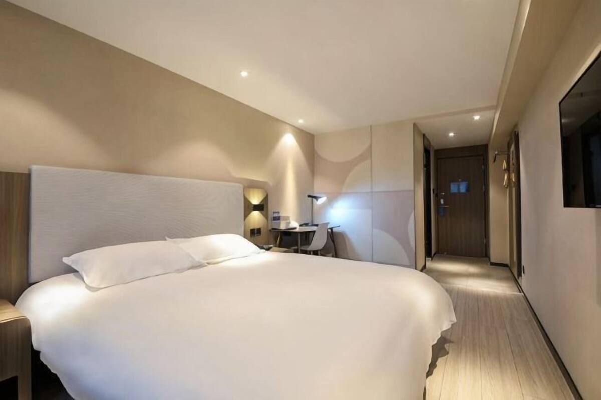 A modern hotel room with a large bed, bedside table, desk, and a view of the hallway.