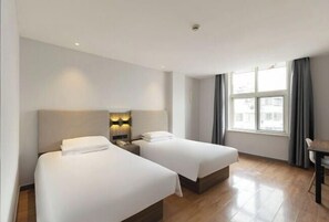 A hotel room with two beds, a desk, a window with curtains, and a city view.