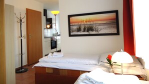 A hotel room with a bed, a bedside table with a lamp, a painting of a sunset, a coat rack, and an adjacent kitchen area. - Uns Wiek-Hus (Mönchgut-Granitz)