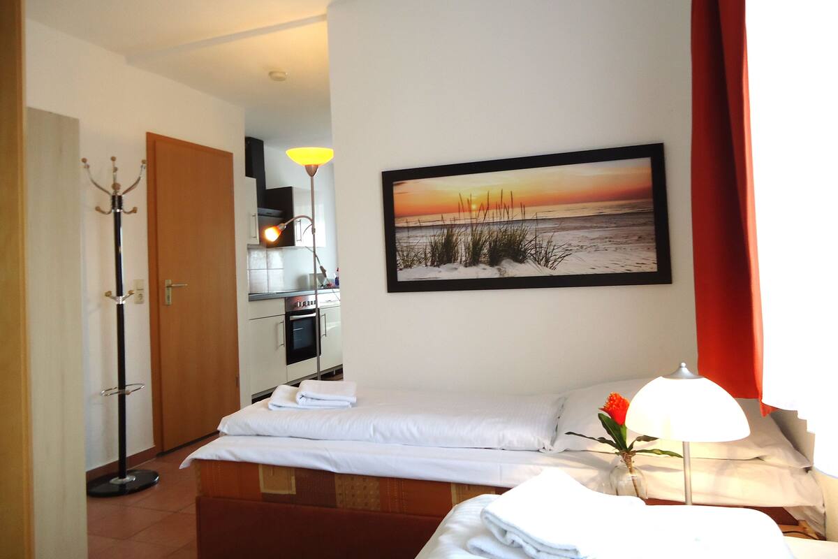 A hotel room with a bed, a bedside table with a lamp, a painting of a sunset, a coat rack, and an adjacent kitchen area.
