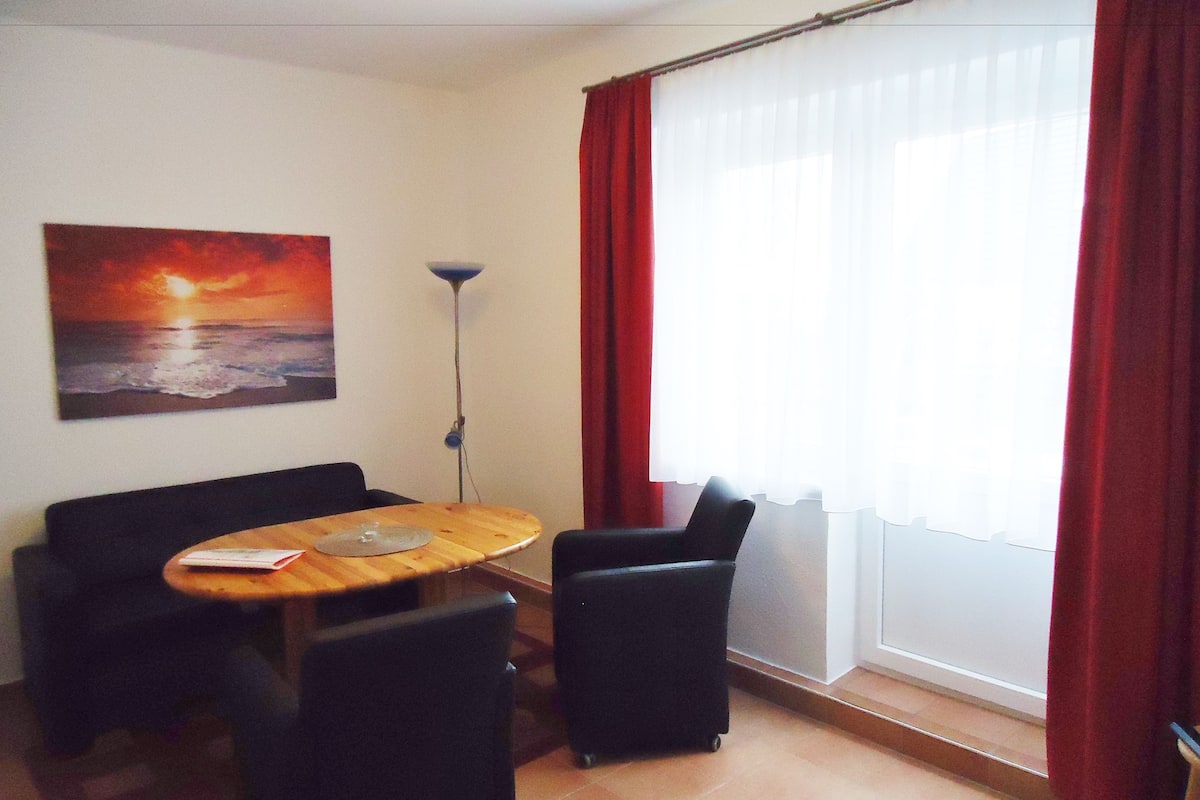 A room with a wooden table, two black chairs, a sofa, a floor lamp, and a large window with red curtains.