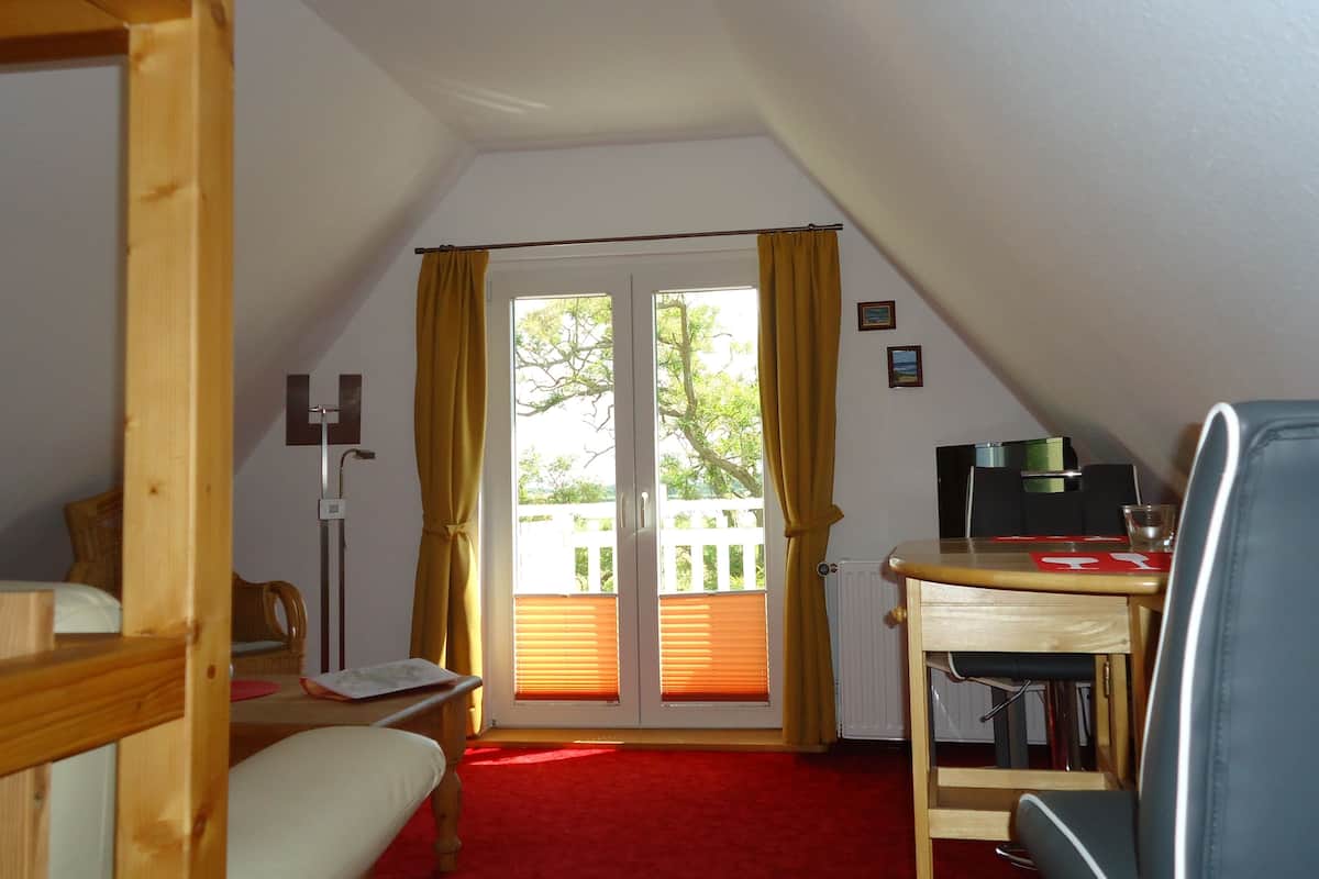 A room with a wooden desk, a chair, a bed, and a red carpet. There is a window with yellow curtains and a view of trees outside.