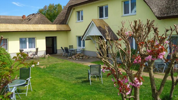 A two-story building with a thatched roof and a light yellow exterior. There are outdoor seating areas with chairs and tables, and a small garden with flowering plants. - Uns Wiek-Hus (Mönchgut-Granitz)