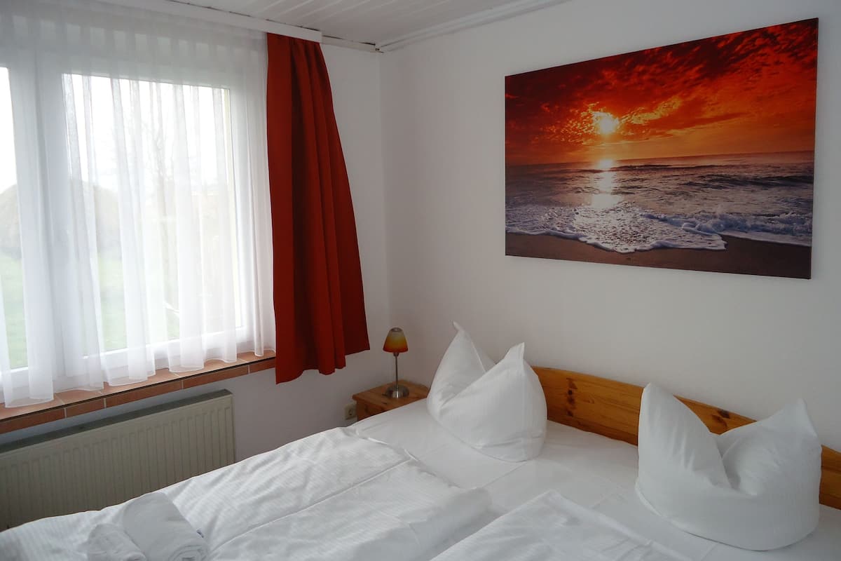 A bedroom with a bed, white bedding, a nightstand with a lamp, a window with sheer curtains and a red curtain, and a sunset painting on the wall.