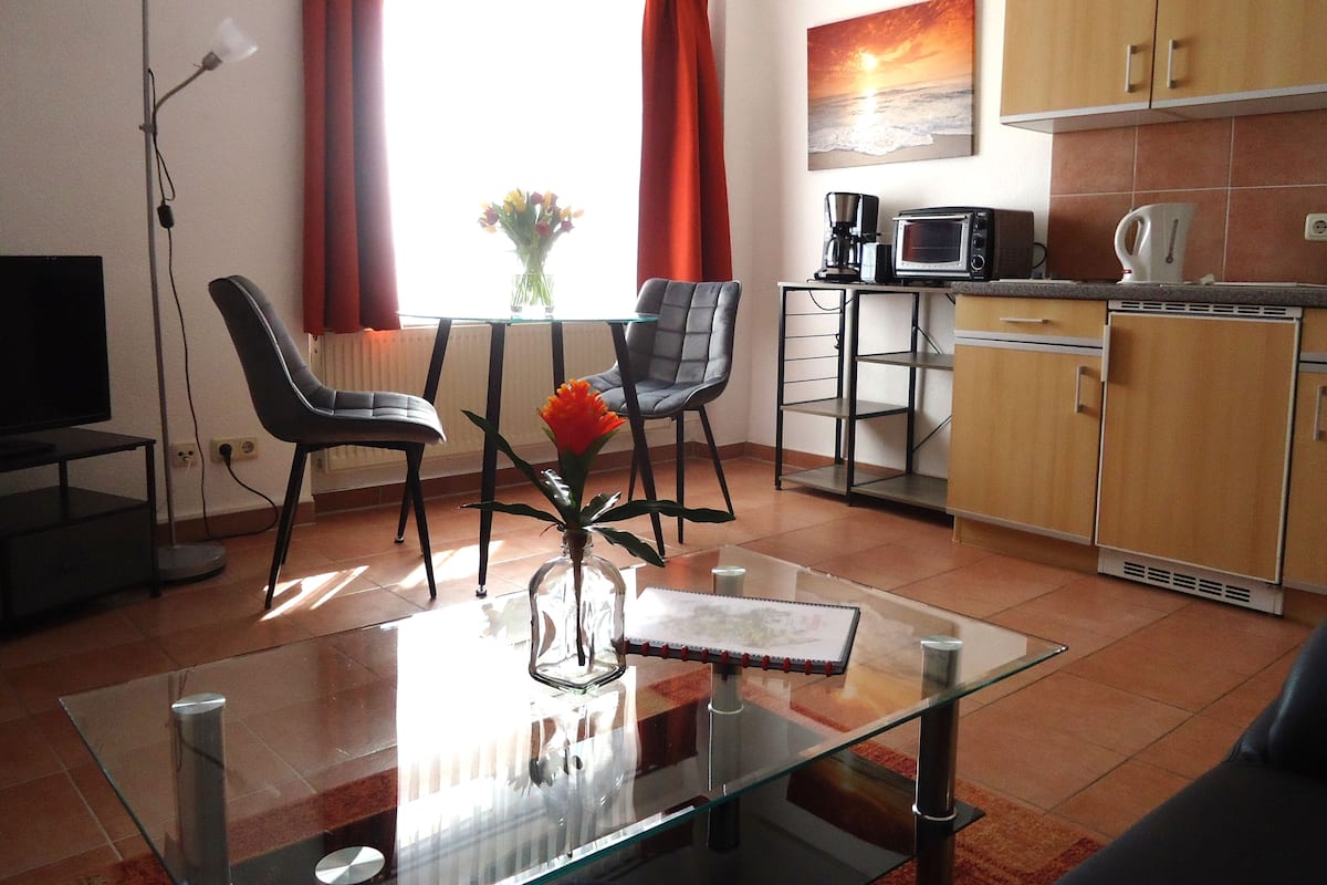 A modern living room with a glass coffee table, two chairs, a TV, and a dining area with a small table and chairs.