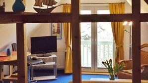 A room with a wooden-framed window, a television on a stand, a chair, and a table with a vase of flowers. - Uns Wiek-Hus (Mönchgut-Granitz)