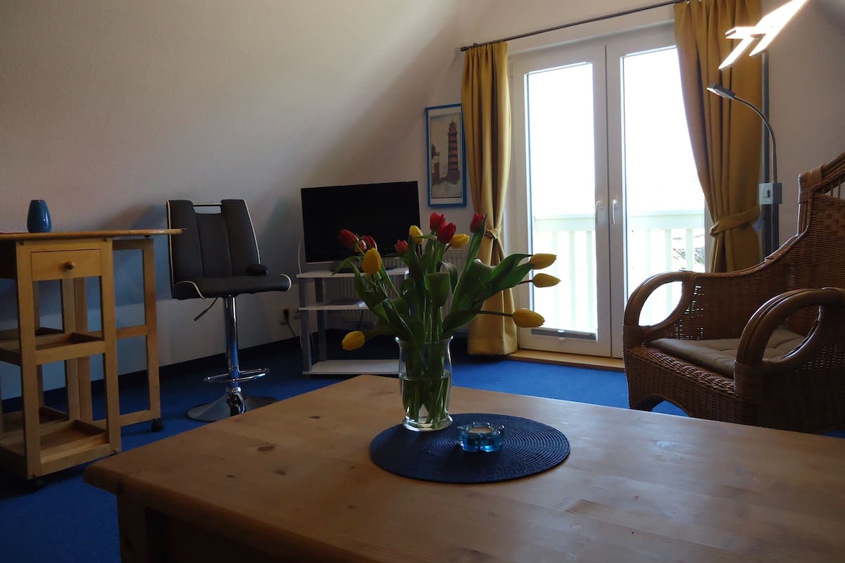 A room with a wooden table, a TV, a chair, and a vase with flowers.