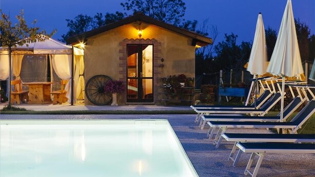Un pequeño edificio con puerta de vidrio, piscina con sillas de descanso y sombrillas.