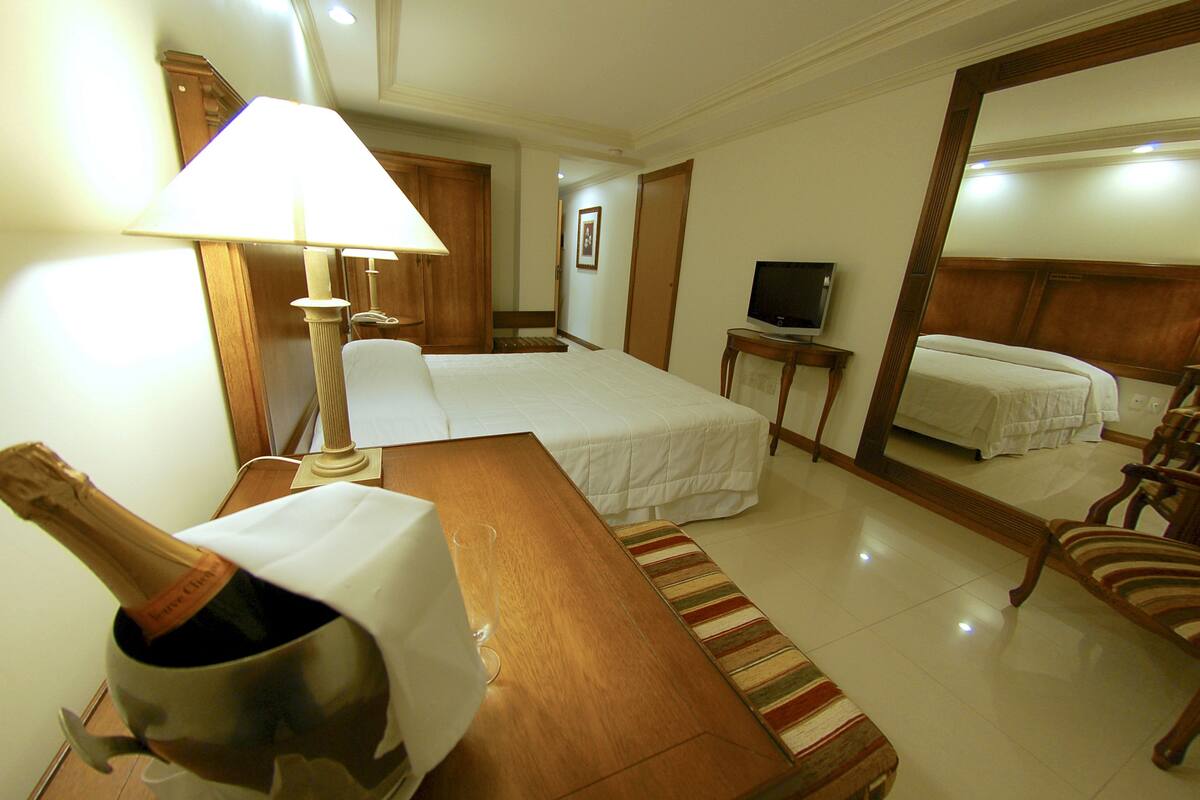Una habitación de hotel con una cama, una mesita de noche con una botella de champán, una lámpara, un televisor y un espejo.