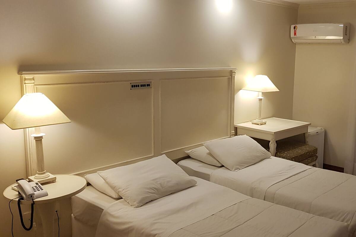 Una habitación de hotel con dos camas, un escritorio, una silla, una lámpara y aire acondicionado.