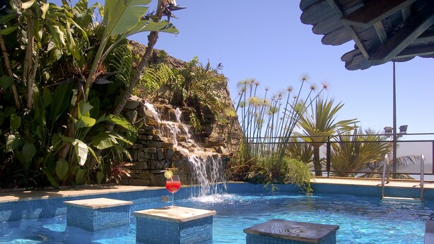 A swimming pool with a waterfall feature, surrounded by lush tropical plants and a stone wall. - Hotel Villa do Mar (Balneário Camboriú)