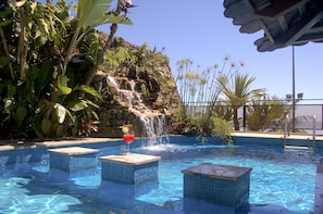 A swimming pool with a waterfall feature, surrounded by lush tropical plants and a stone wall.