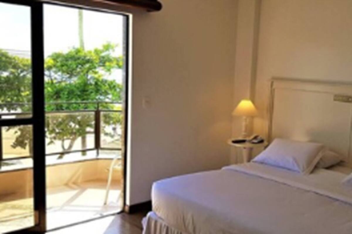 Habitación de hotel con una cama grande, una mesita de noche con lámpara y vistas a zonas verdes por la ventana.