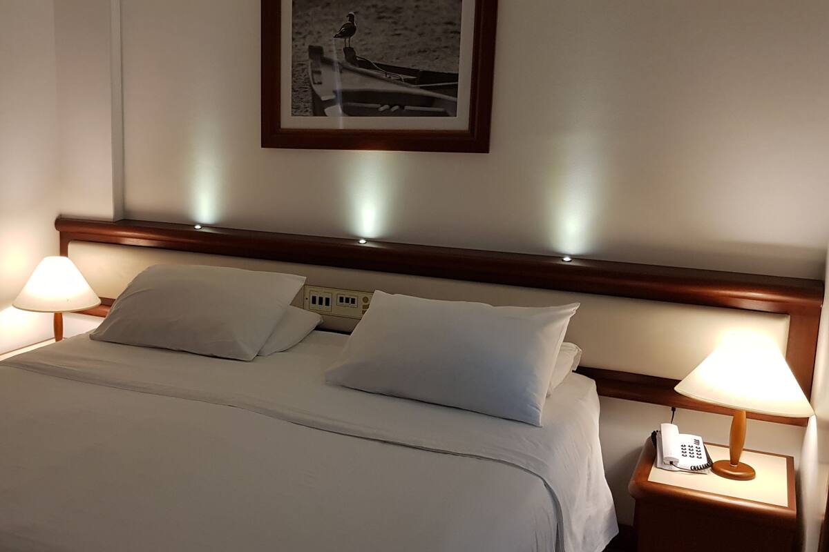 Una cama bien tendida con sábanas blancas, dos lámparas de noche, una mesita con teléfono y un cuadro en la pared.