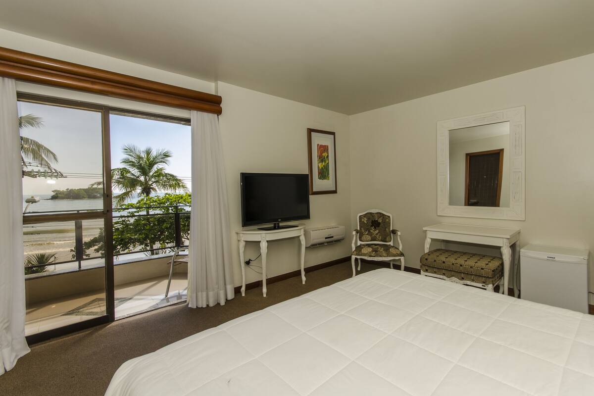 Habitación de hotel con cama, televisión, una silla, una mesita y vistas a la playa.