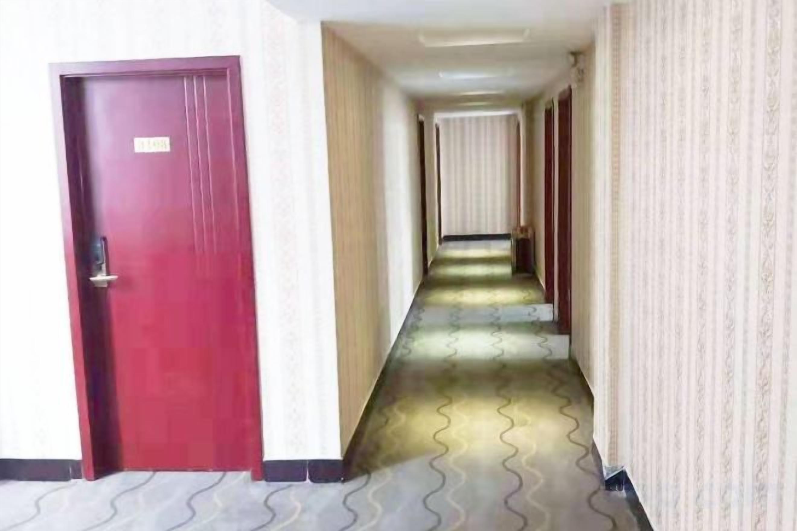 A hotel hallway with a red door, patterned carpet, and a long corridor with doors at the end.