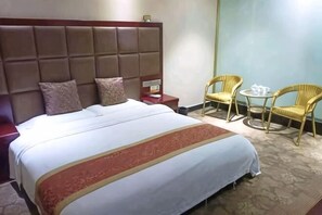 Een hotelkamer met een groot bed, twee kussens, een nachtkastje, een kleine tafel met een stoel en een tweede stoel.