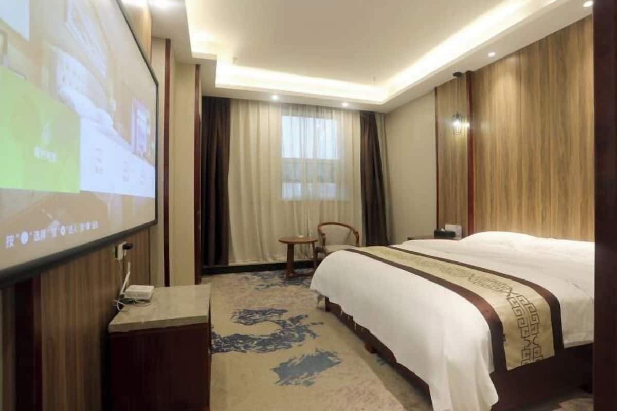 Ein Hotelzimmer mit Bett, Schreibtisch, Stuhl, großem Bildschirm und einem Fenster mit Vorhängen.