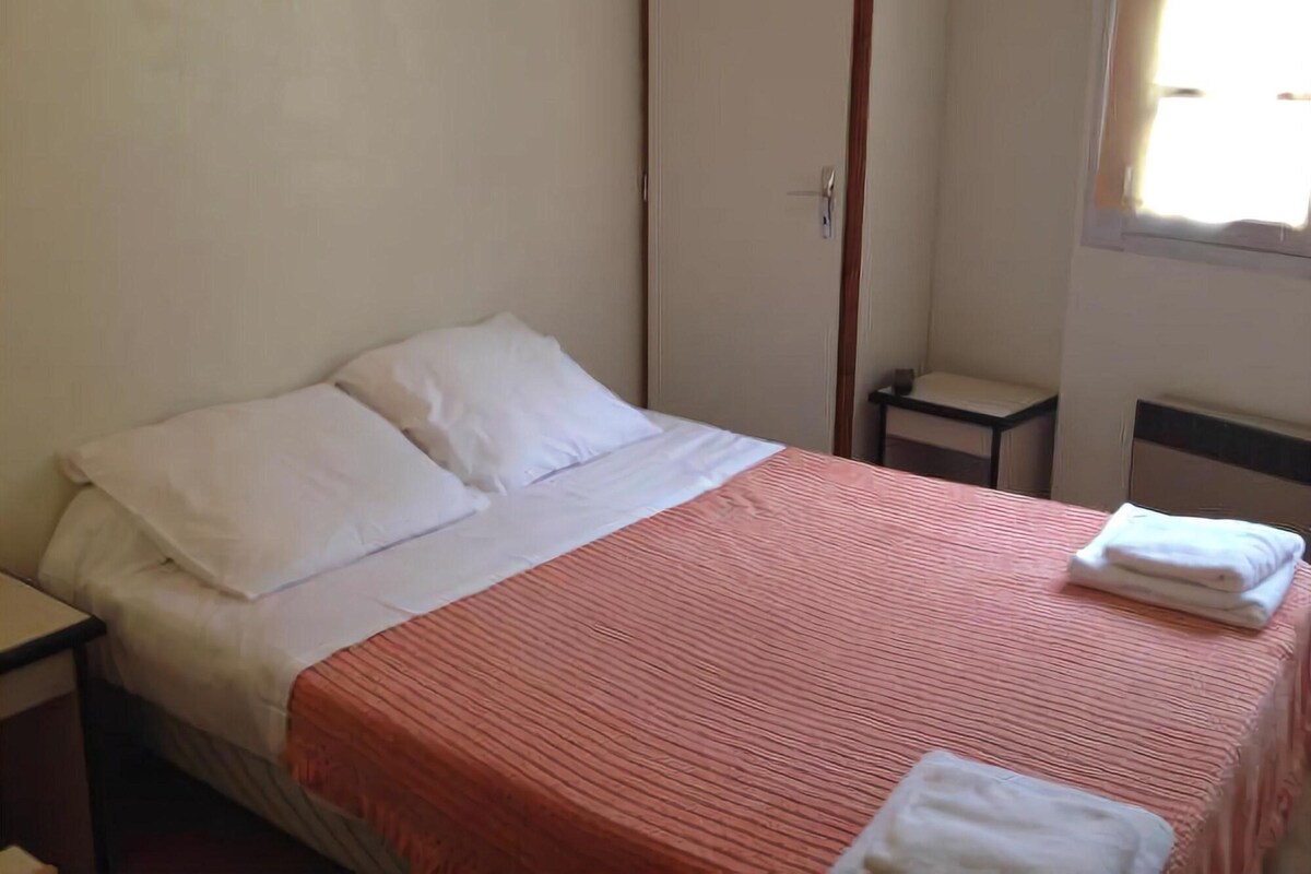 Una cama individual con almohadas blancas y una colcha a rayas rojas, una mesita de noche, una pequeña mesa y una ventana con persianas.