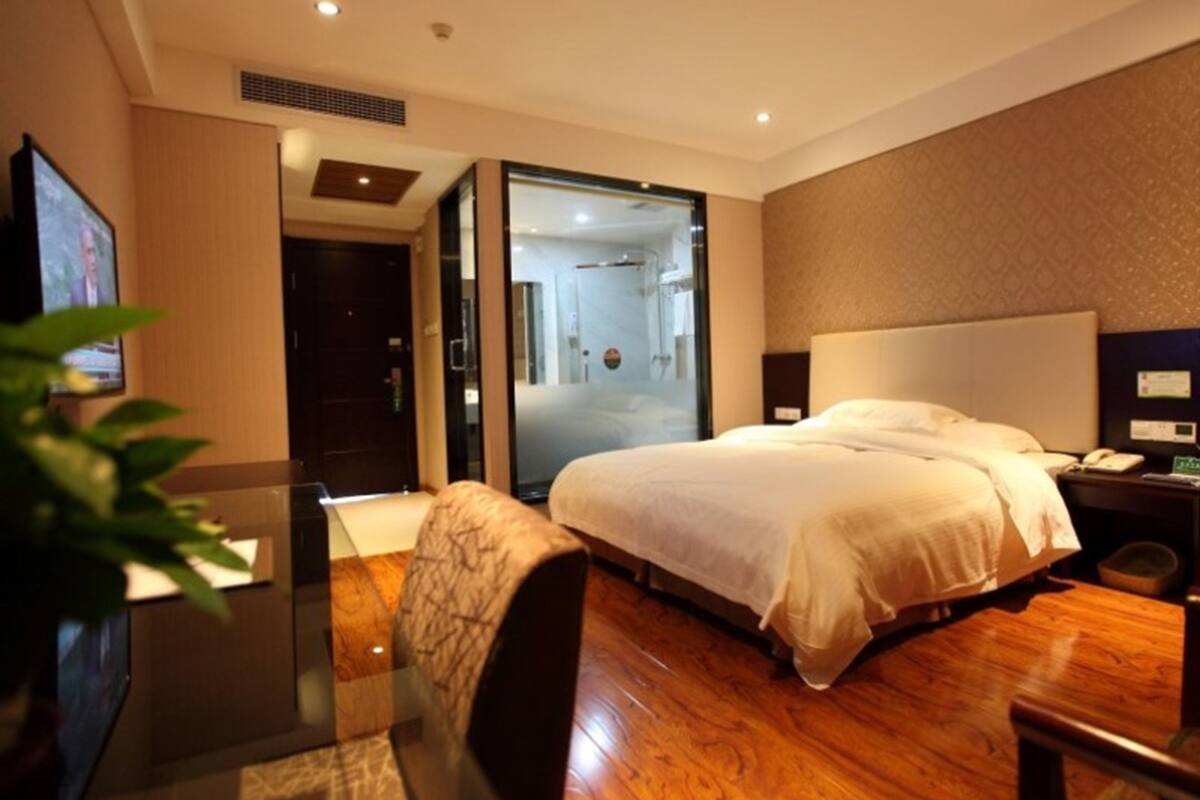 Hotelkamer met een groot bed, een bureau met stoel, een nachtkastje met lamp, en uitzicht op de badkamer door een glazen deur.