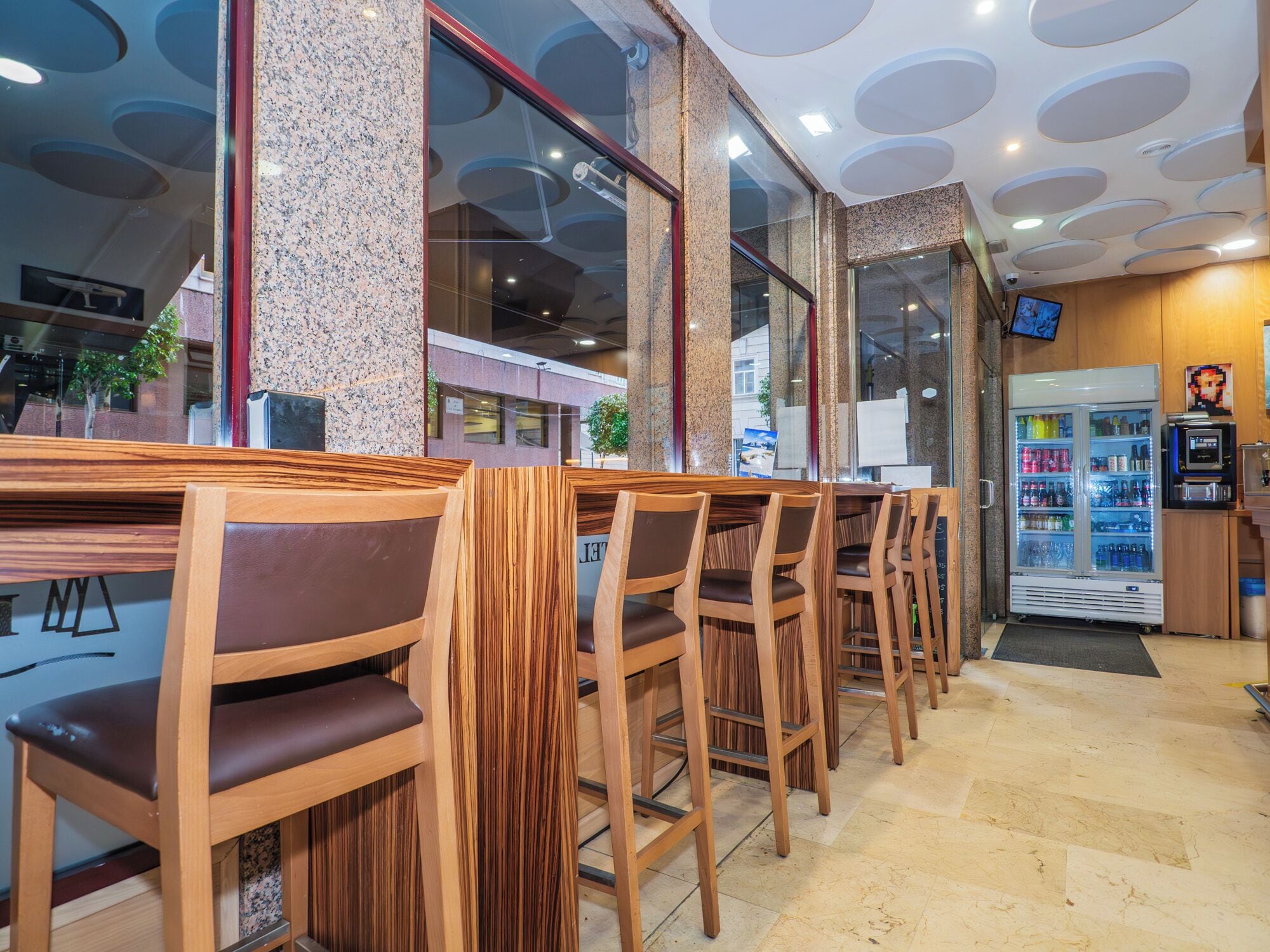 A restaurant interior with wooden bar seating, a refrigerator with beverages, and a television on the wall.
