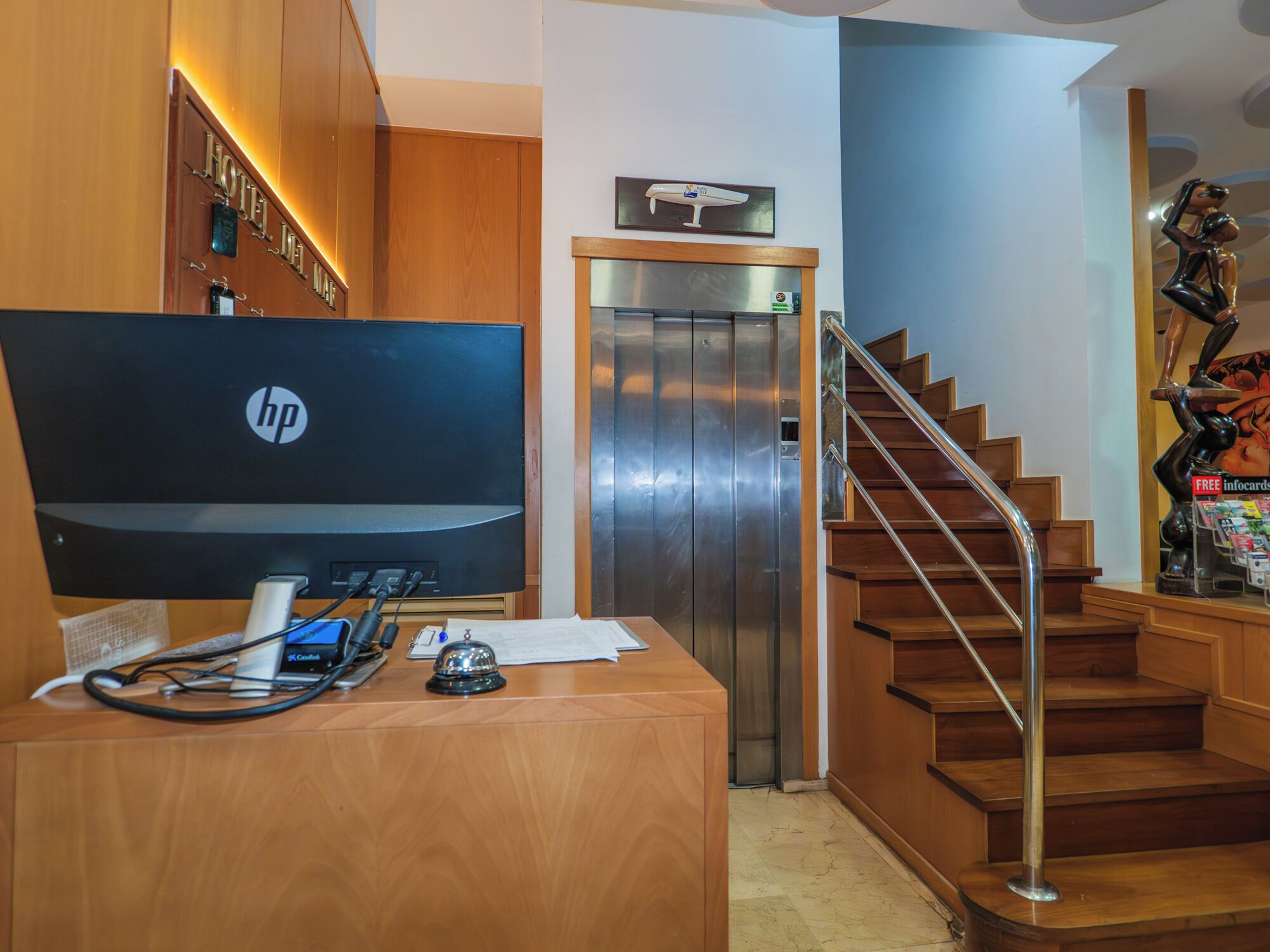 Een receptiebalie van een hotel met een HP monitor, een telefoon en een bel. Er is een lift en een trap met een metalen leuning.