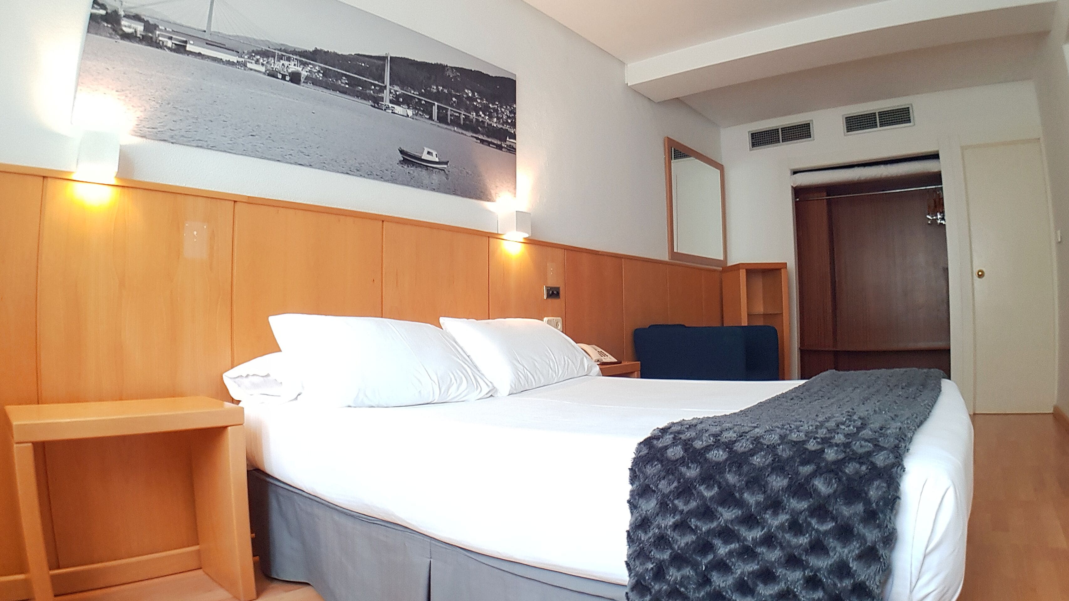 A hotel room with a bed, a nightstand, a chair, a wardrobe, and a wall-mounted picture of a bridge.