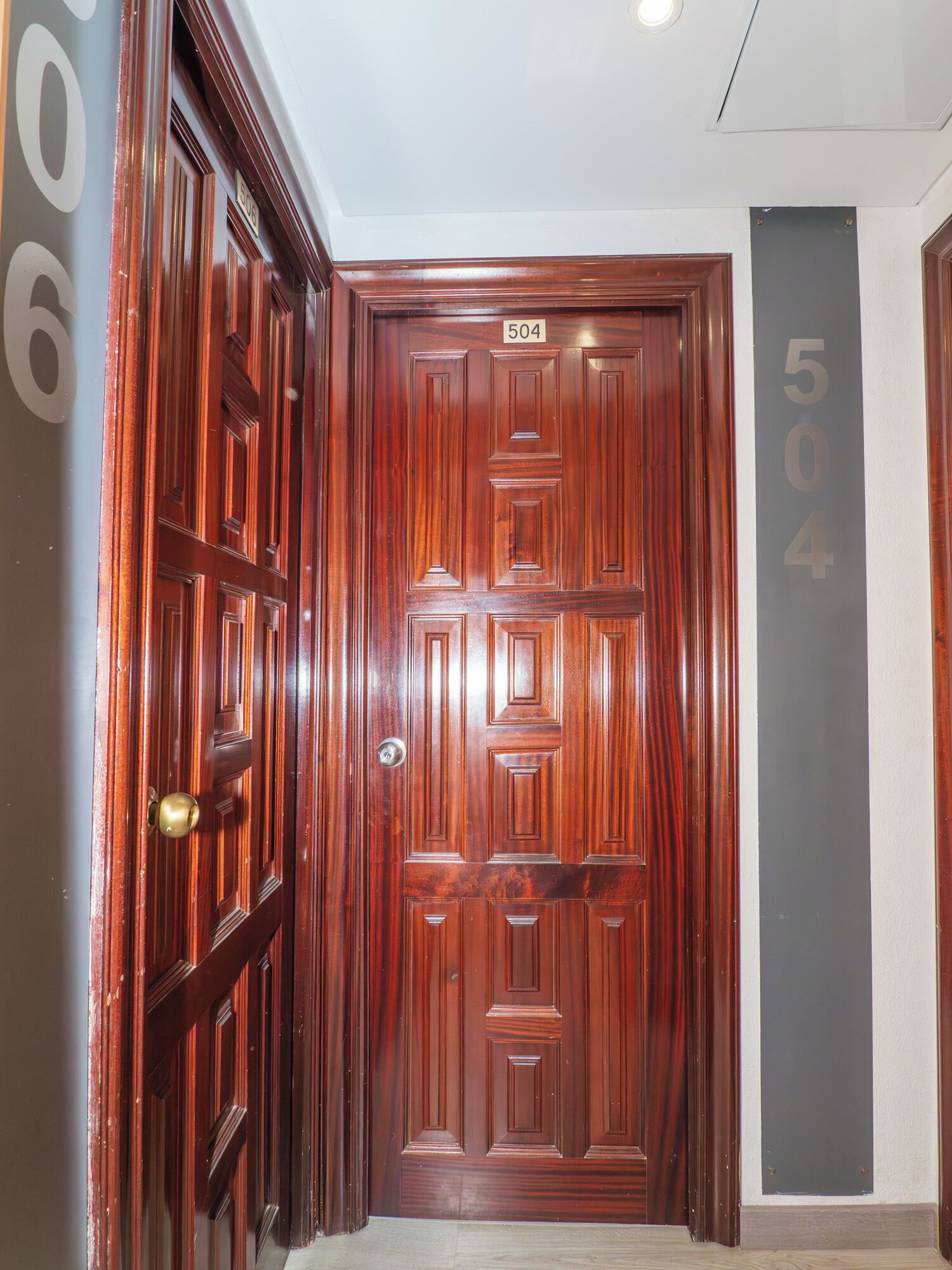 Una puerta de madera con una placa de número, un pasillo con números en la pared y un suelo con baldosas de color claro.