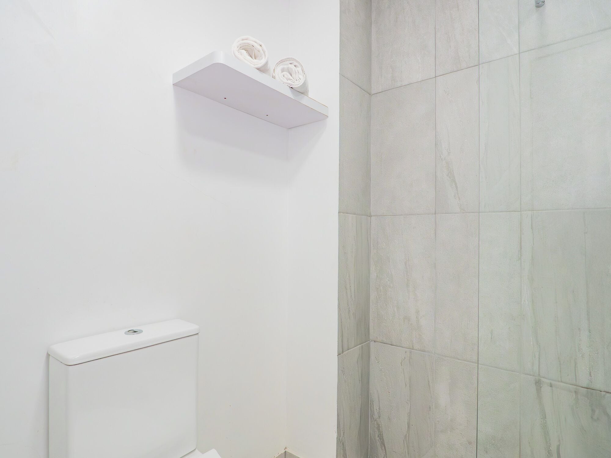 Une salle de bain avec des toilettes, une étagère blanche avec des serviettes et un mur recouvert de carreaux.