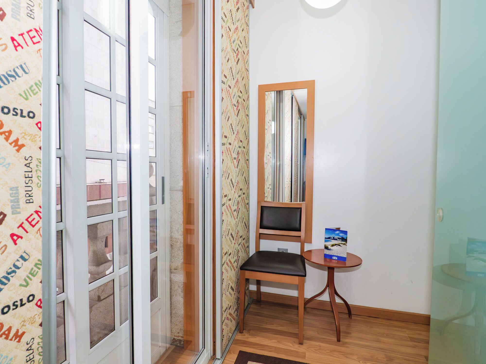 Una habitación con una puerta corrediza de vidrio, una silla de madera, una mesita con un libro y un espejo en la pared.