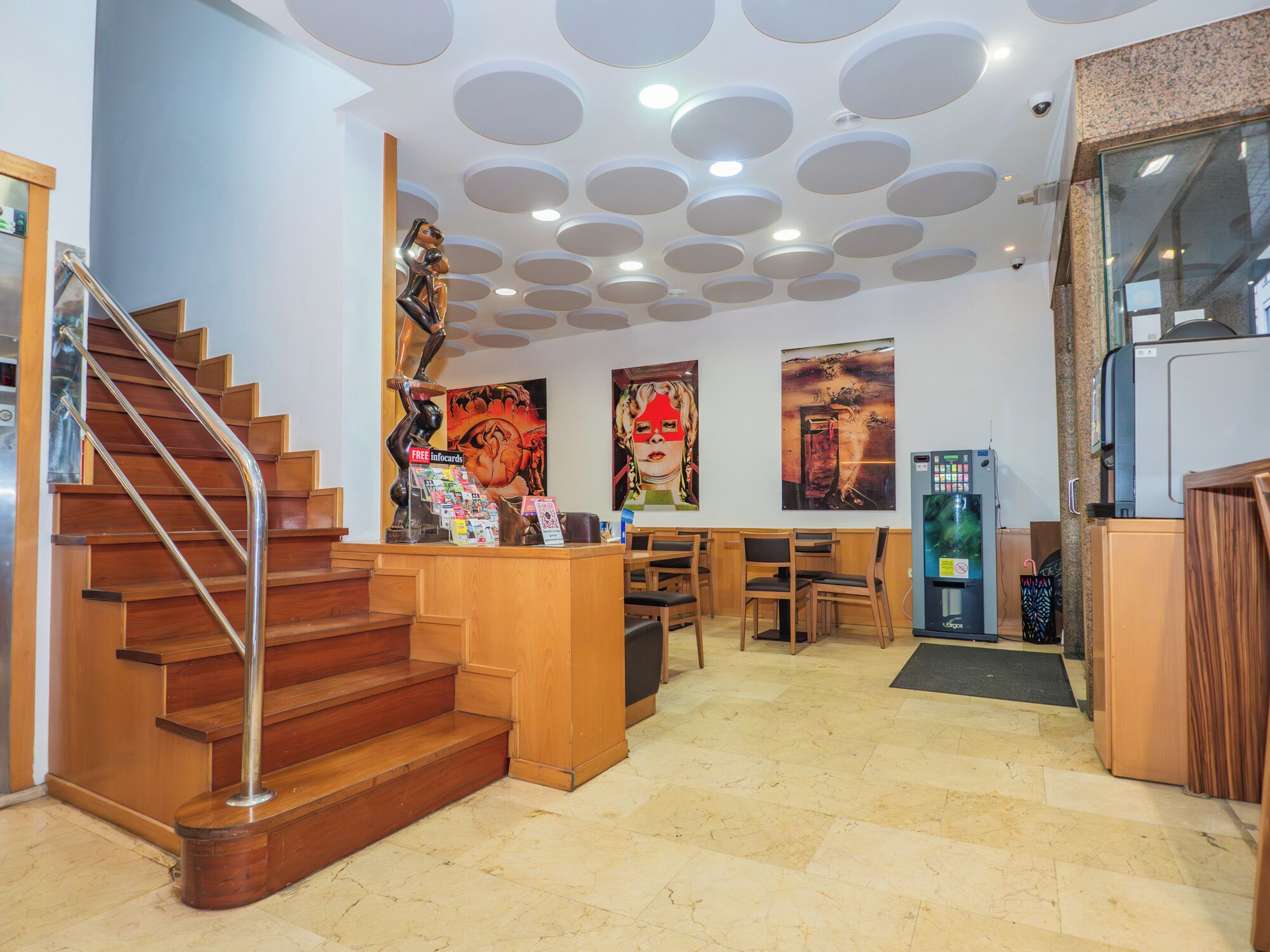 Un hall d’hôtel avec un escalier en bois, un comptoir d’accueil et un mur orné de luminaires circulaires au plafond.