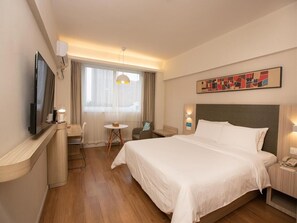 Habitación de hotel con una cama grande, un escritorio con silla, una mesita, televisión y ventana con cortinas.