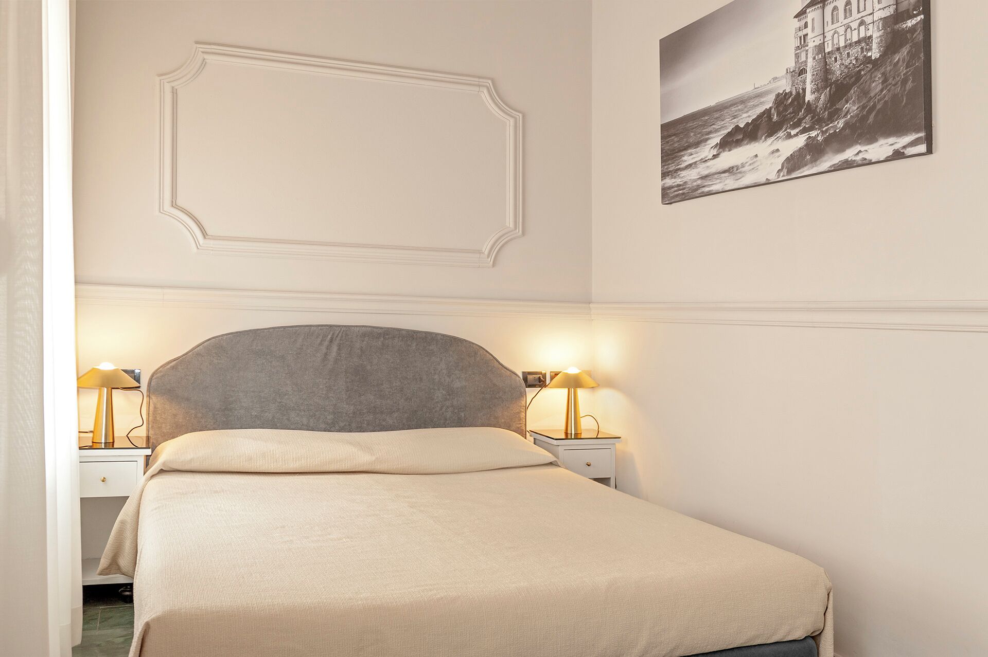 Een netjes opgemaakt bed met een grijze hoofdbord, twee bedlampen, een ingelijste foto aan de muur en decoratieve sierlijsten boven het bed.