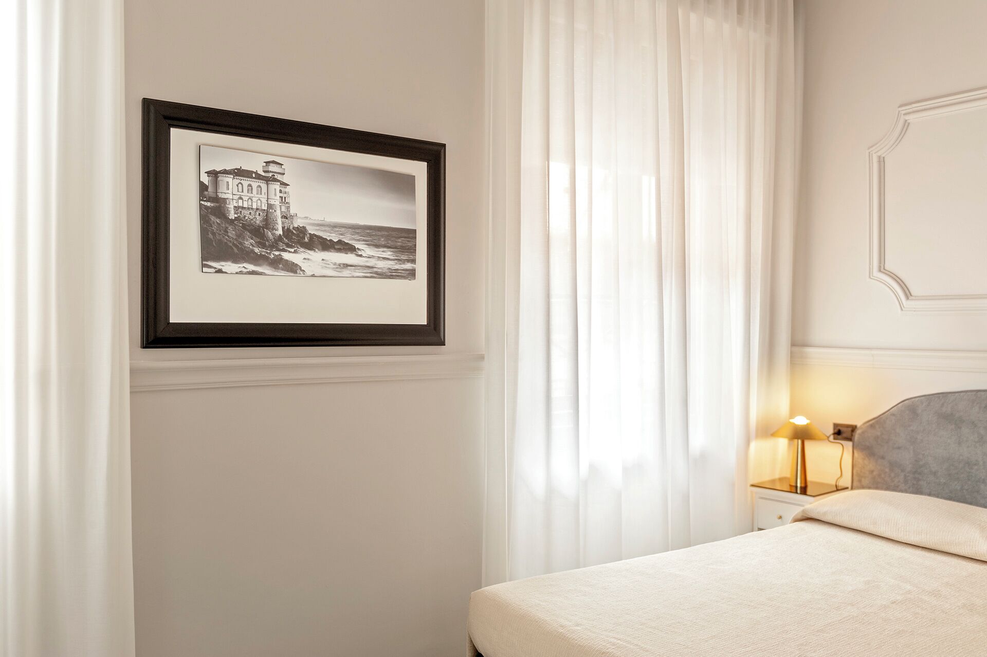 A neatly arranged bedroom with a framed black and white photograph on the wall, a bed with a light-colored bedspread, a bedside lamp, and sheer white curtains.