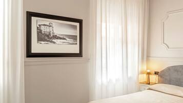 A neatly arranged bedroom with a framed black and white photograph on the wall, a bed with a light-colored bedspread, a bedside lamp, and sheer white curtains.