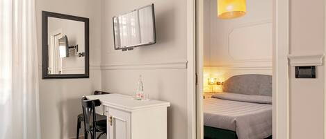 A hotel room with a bed, a desk with a chair, a TV mounted on the wall, and a door leading to another room.