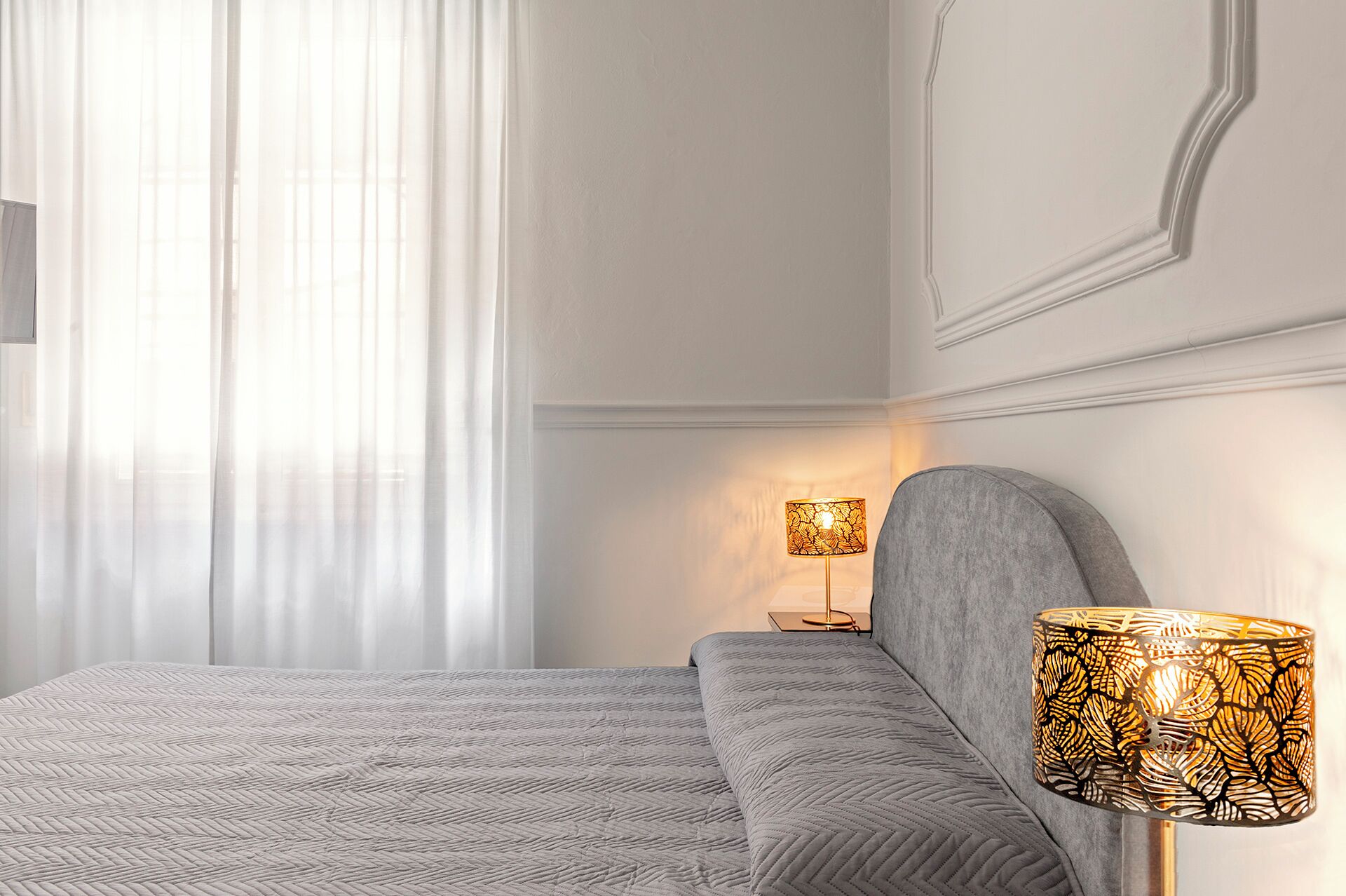 Une chambre d’hôtel avec un lit, une table de chevet, une lampe et une fenêtre avec des rideaux légers.