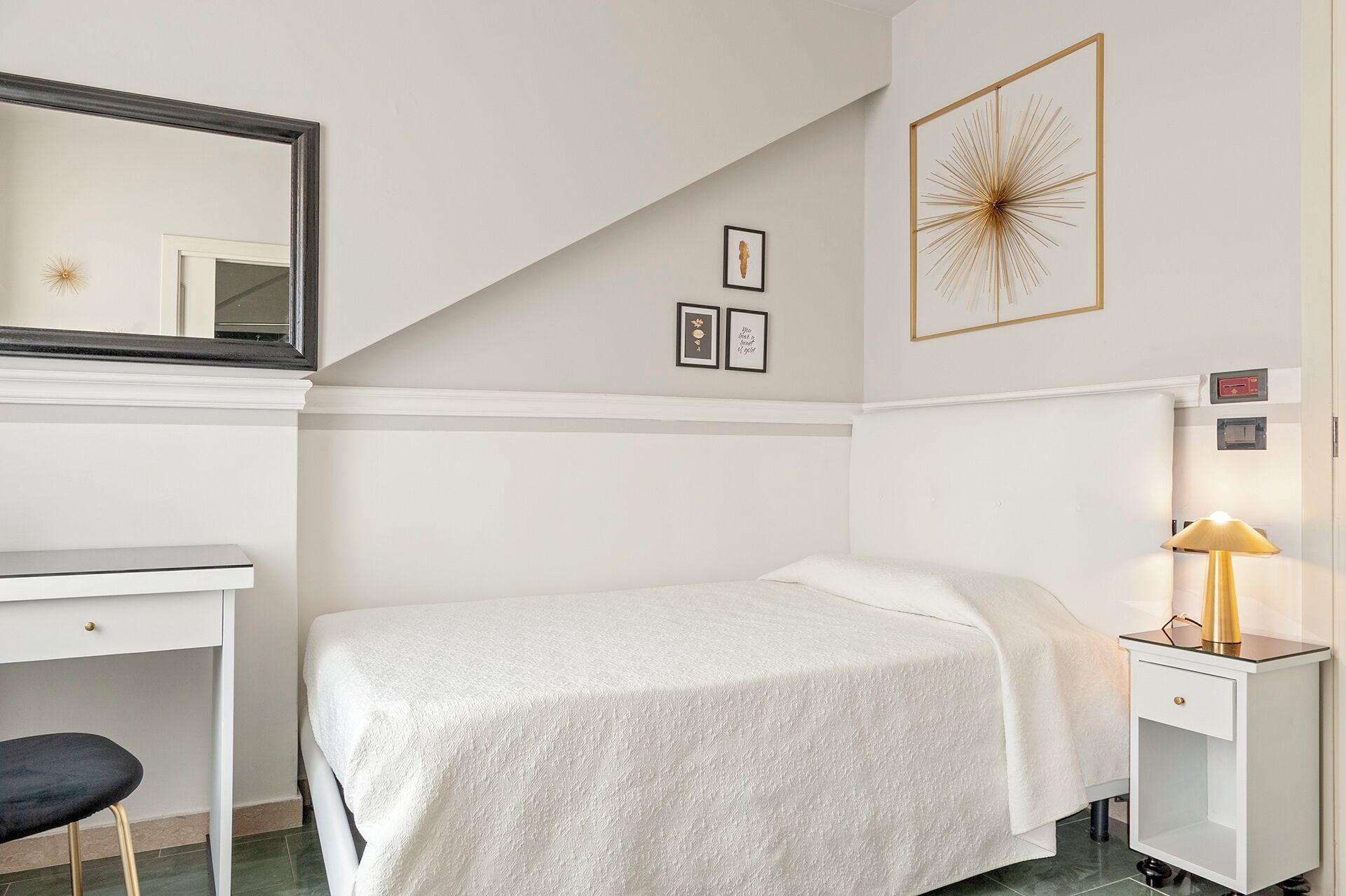 A bedroom with a bed, a desk, a chair, a bedside table with a lamp, and wall art.