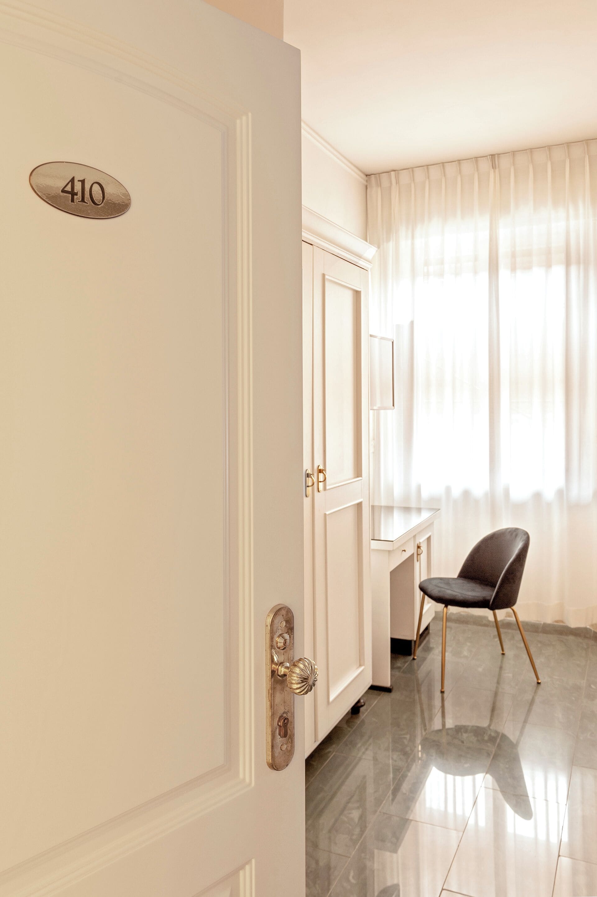 A hotel room with a white door marked "410", a desk with a chair, and a large window with sheer curtains.