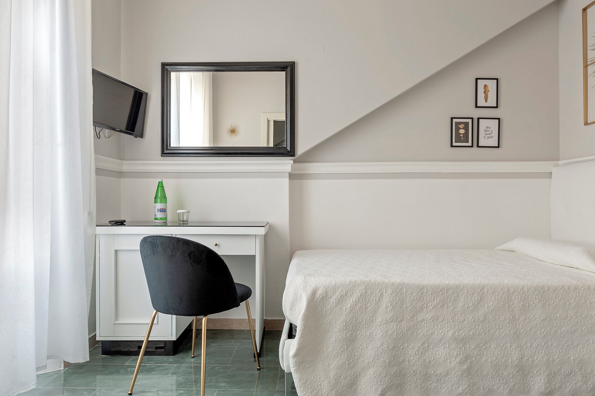 A hotel room with a bed, a desk with a chair, a TV mounted on the wall, and a window with curtains.