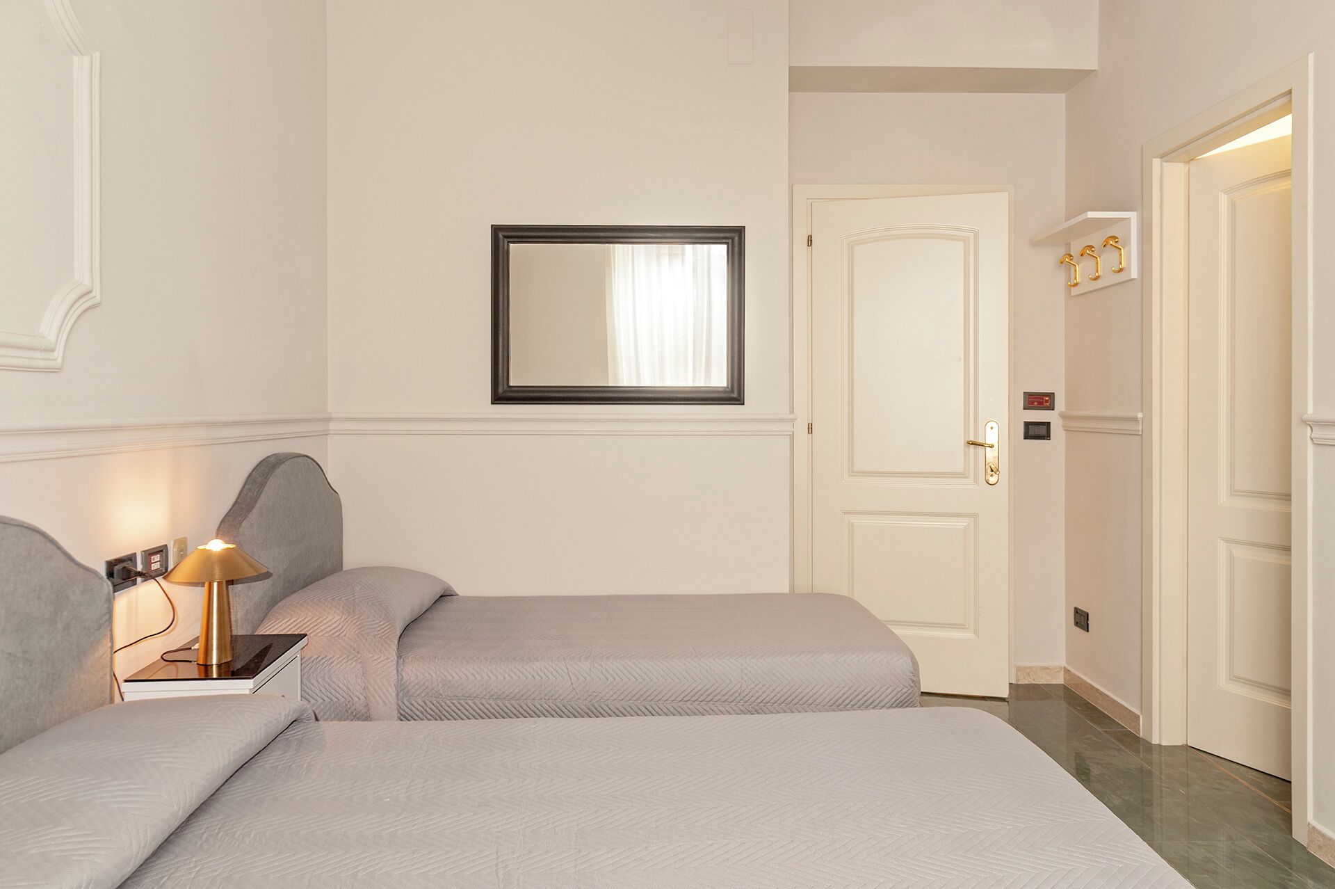 A hotel room with two beds, a bedside table with a lamp, a mirror, and a door with a light switch.