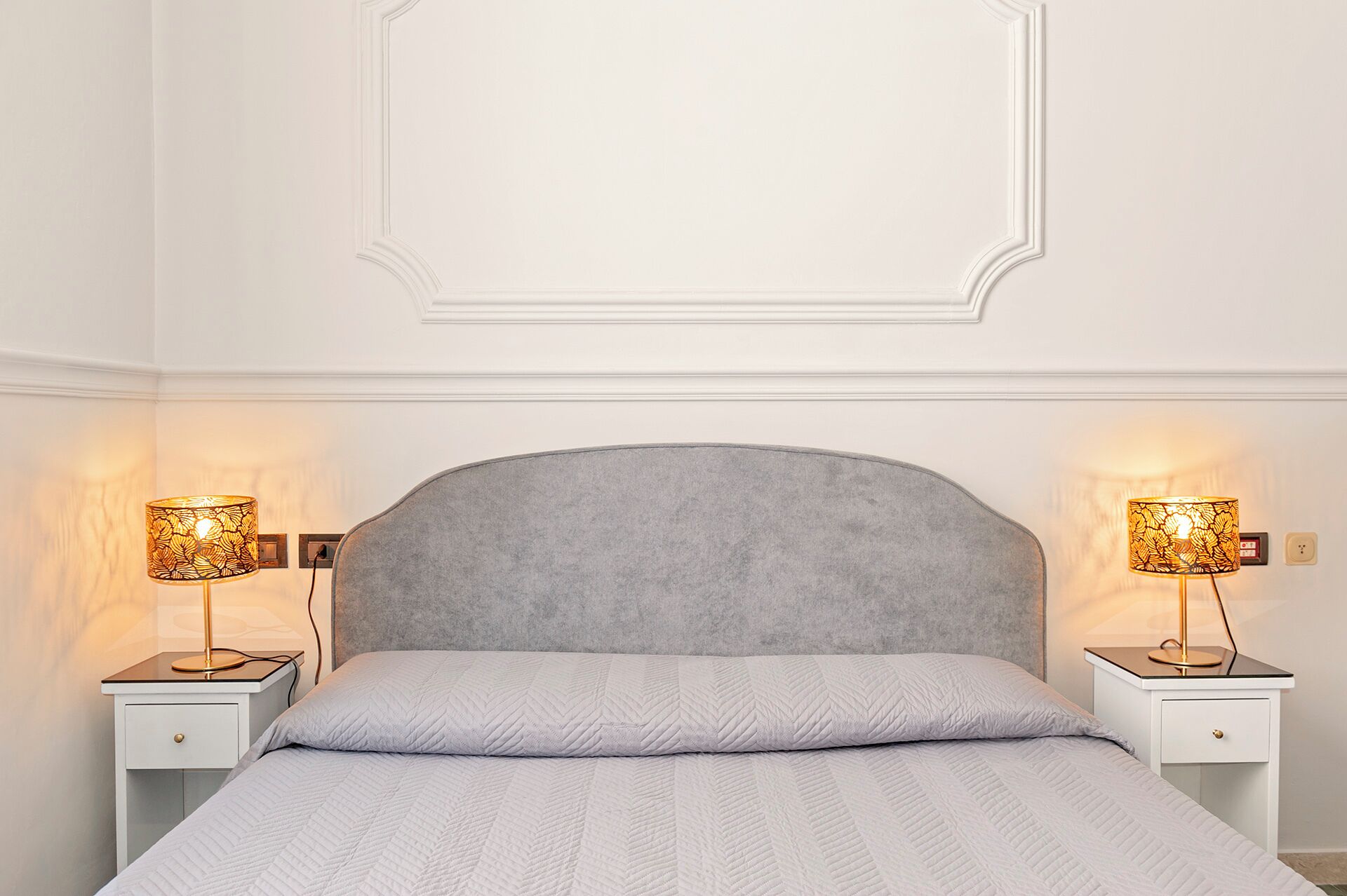 Une chambre à coucher avec une tête de lit rembourrée de couleur grise, une couvre-lit blanche matelassée et deux lampes de chevet dotées d’abat-jour à motifs.