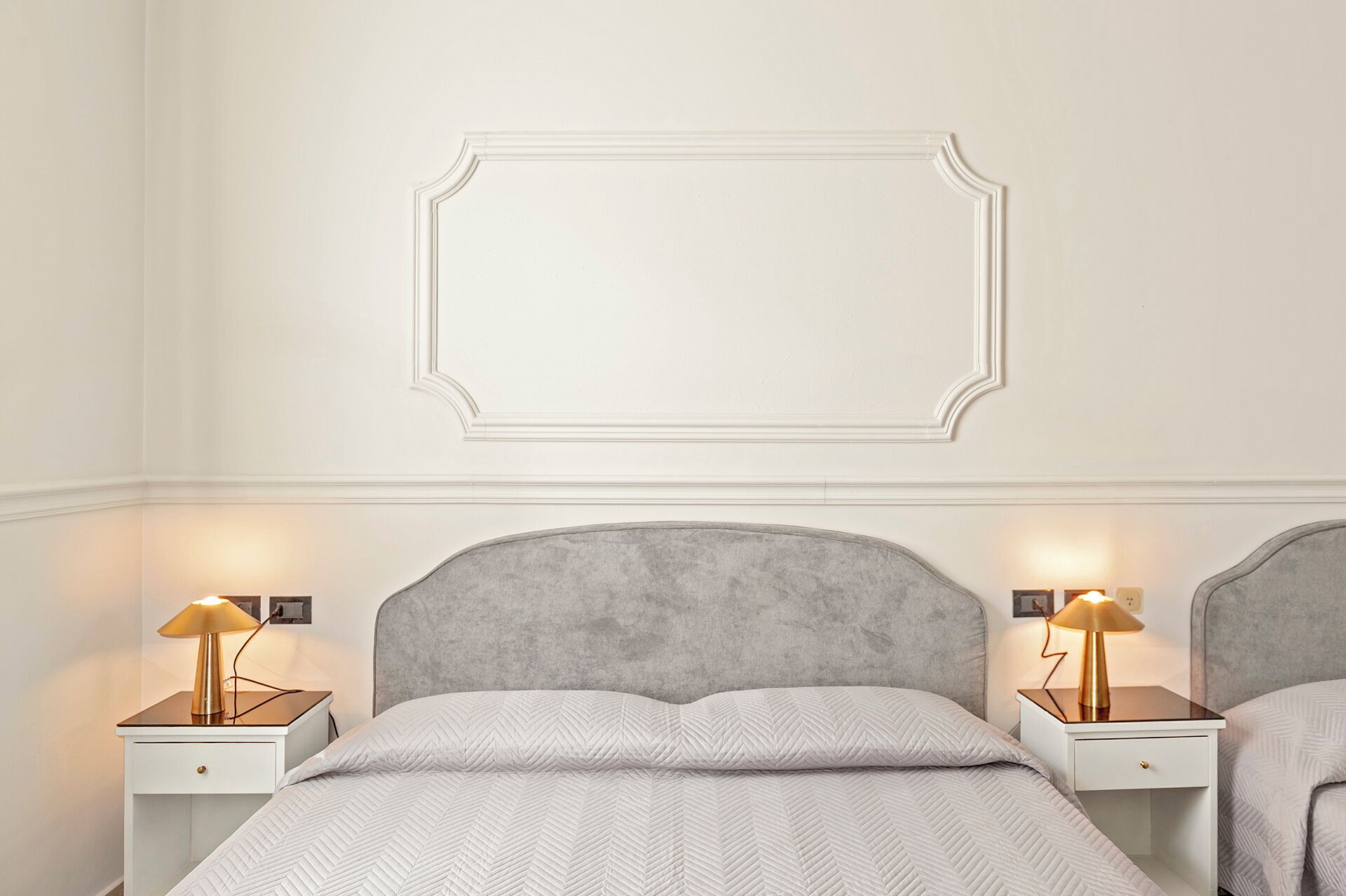 A bedroom with a grey upholstered headboard, a white quilted bedspread, and two bedside tables with lamps.