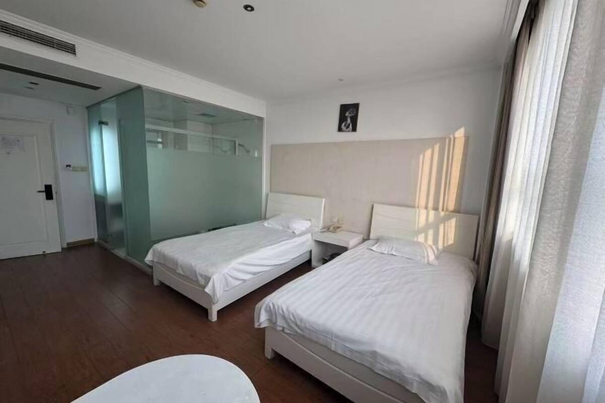 A hotel room with two beds, a glass partition, a white door, and a small table.