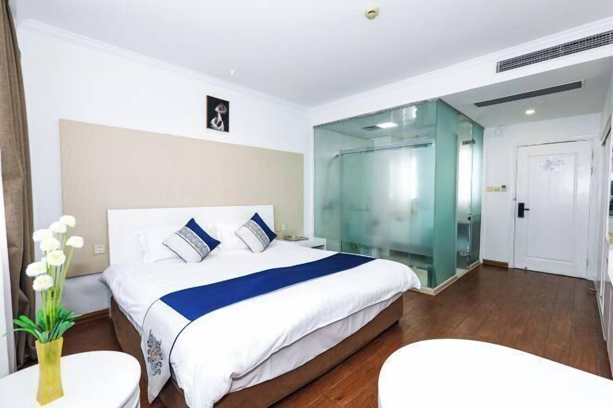 A hotel room with a bed, a glass partition, a white door, and a small table with a vase.