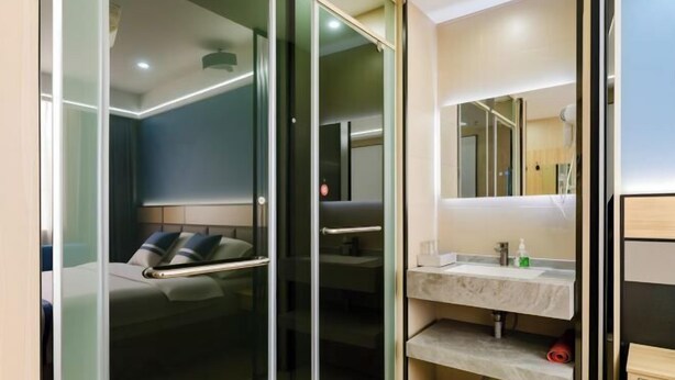 Una habitación de hotel moderna con una puerta de vidrio que da a un dormitorio, un tocador de mármol y un pequeño espacio de almacenamiento.