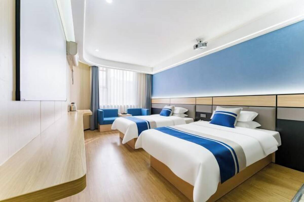 Une chambre d’hôtel avec deux lits, un bureau en bois aux lignes courbes, une grande fenêtre avec des rideaux et un mur d’accent bleu.