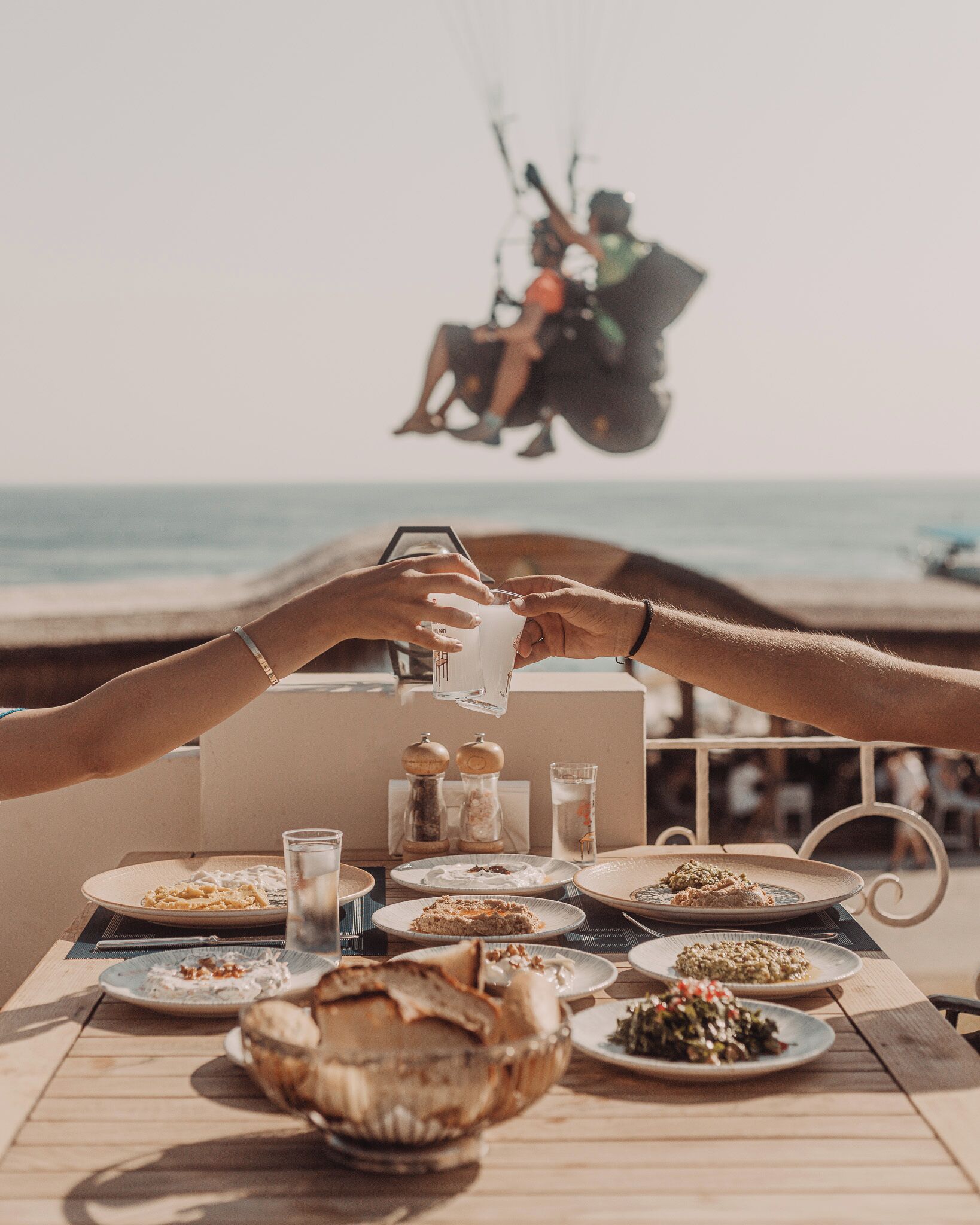 A table set for a meal with various dishes and glasses, overlooking a beach with people in the background.