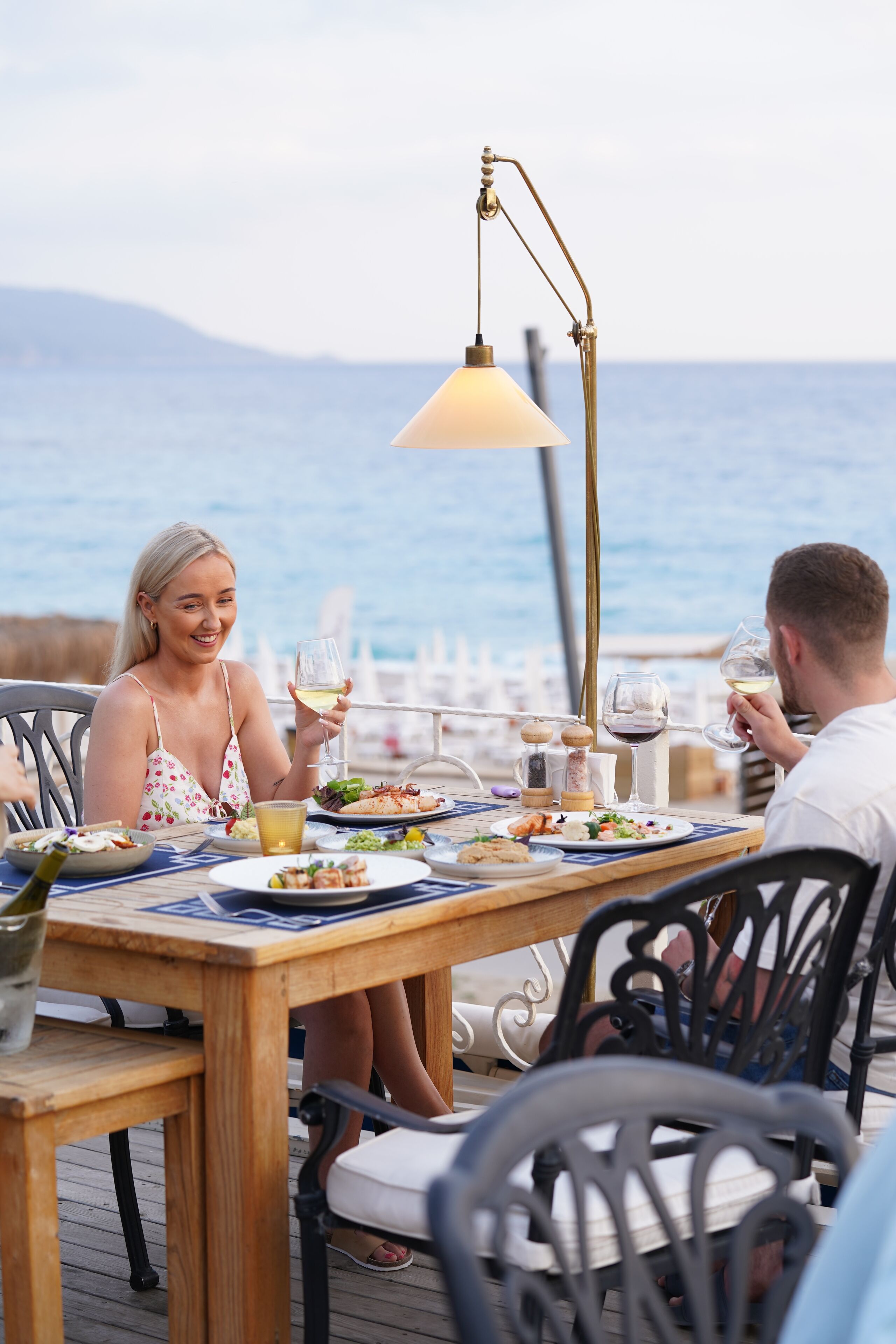 A wooden table with a lamp, plates of food, and glasses of wine.
