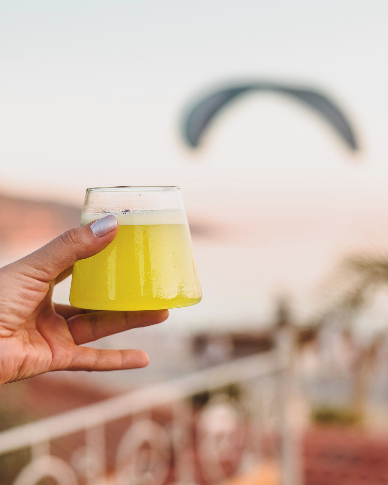 Una mano che tiene un bicchiere di liquido giallo, con sullo sfondo sfocato una struttura curva.
